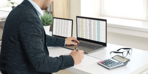 Muž pracujúci s účtovníckymi údajmi na dvoch monitoroch, s kalkulačkou a poznámkami – symbolizuje digitalizované a moderné účtovníctvo.